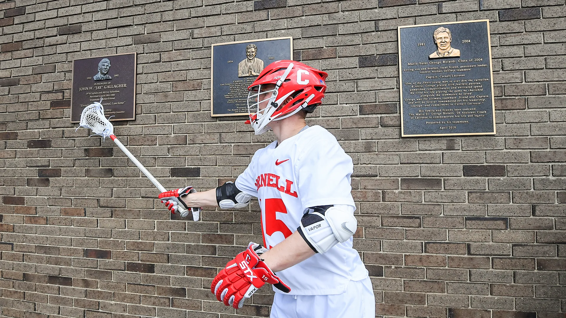 CJ Kirst pays homage to late Cornell greats Jay Gallagher, Eamon McEneaney and George Boiardi by tapping their plaques with his stick at Schoellkopf Field.