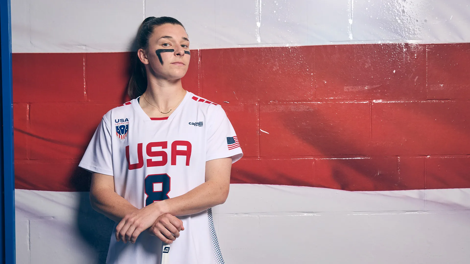 Charlotte North stands against a red, white and blue wall in the locker room at USA Lacrosse's Tierney Field. 