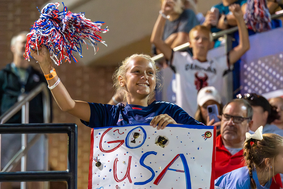 Young girl waving a flag and holding up a USA sign in the stands at 2025 Atlas Cup